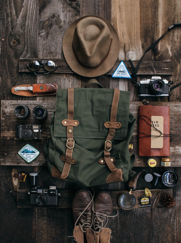 Overhead of vintage photo cameras near backpack and knife near bump and hat near lens near notepad and sunglasses on wooden surface