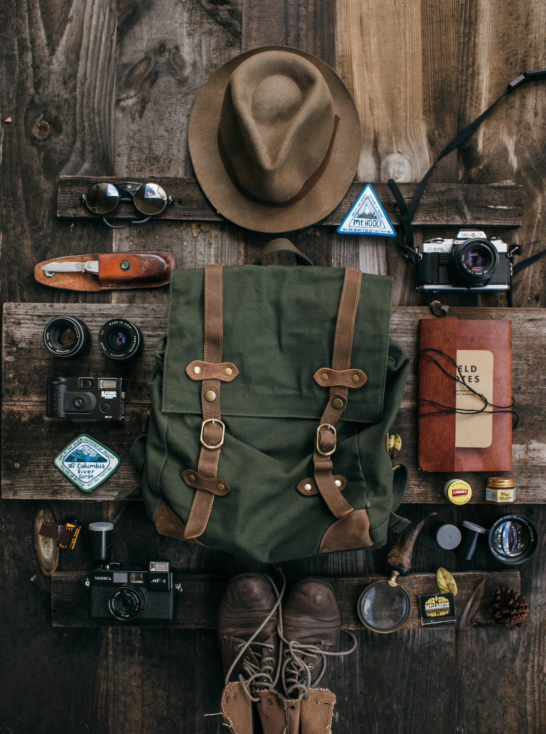 Overhead of vintage photo cameras near backpack and knife near bump and hat near lens near notepad and sunglasses on wooden surface